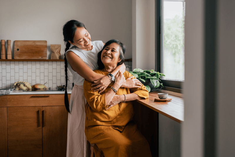 A Happy Beautiful Woman Hugging Her Mother While She Is Sitting In The Kitchen And Drinking Tea - stock photo
A smiling Asian woman enjoying drinking tea while her loving daughter is embracing her.